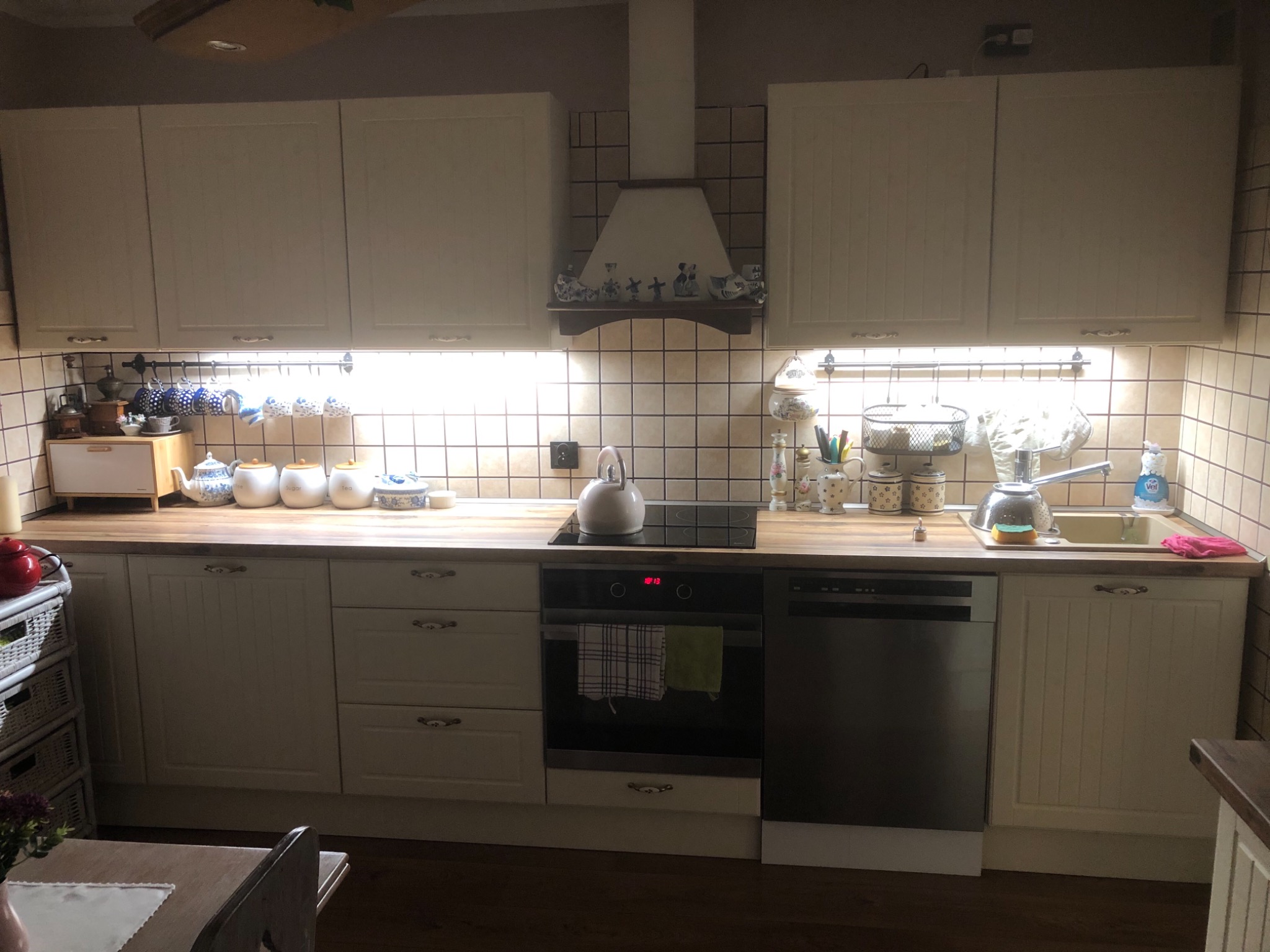 Jasna kuchnia w stylu rustykalnym z beżowymi płytkami, drewnianym blatem, białymi szafkami i okapem ozdobionym holenderskimi figurkami.