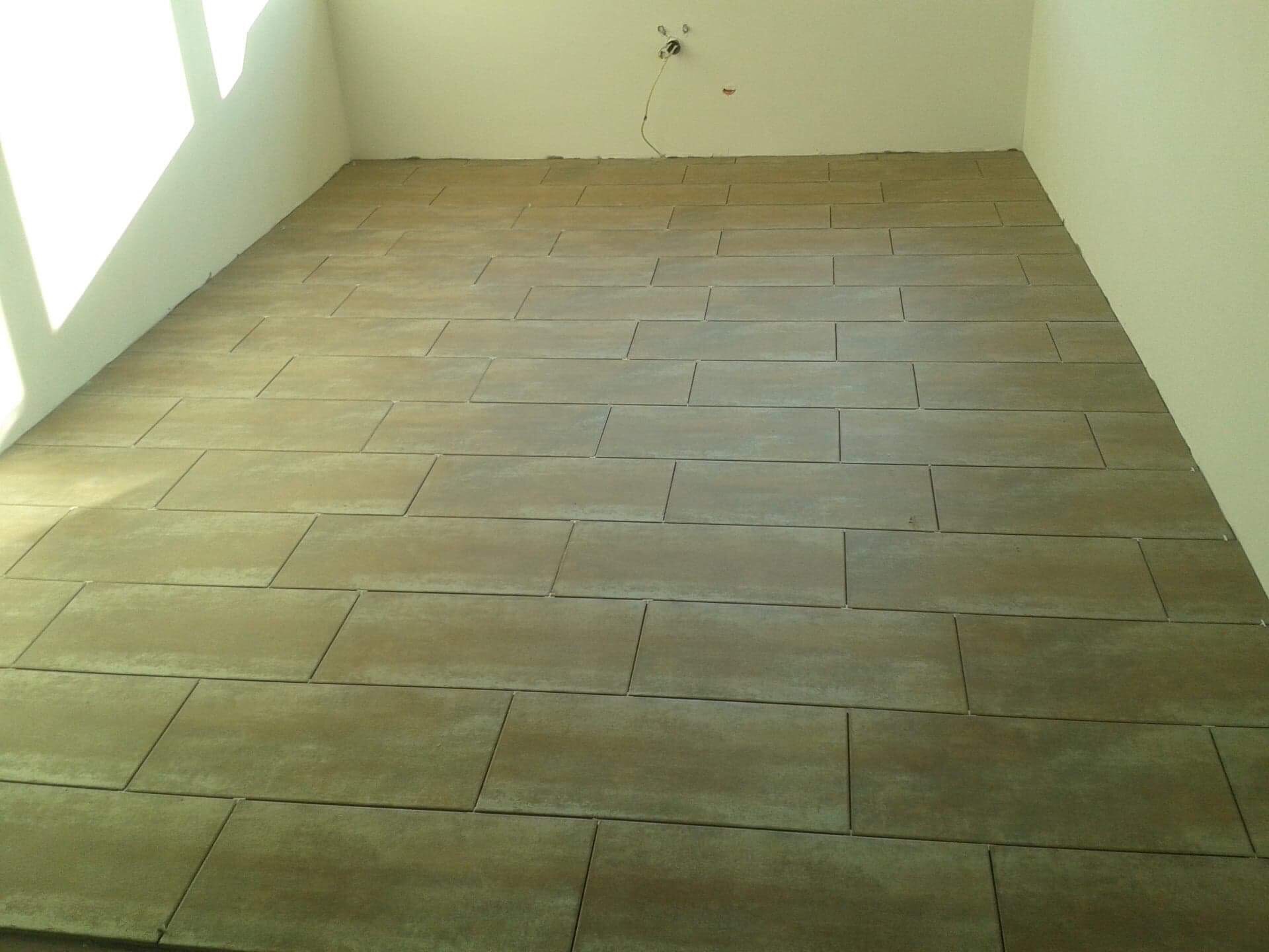 Ułożona podłoga z prostokątnych, matowych płytek w odcieniach beżu i szarości w pustym pomieszczeniu z białymi ścianami, widoczne łączenia fug.