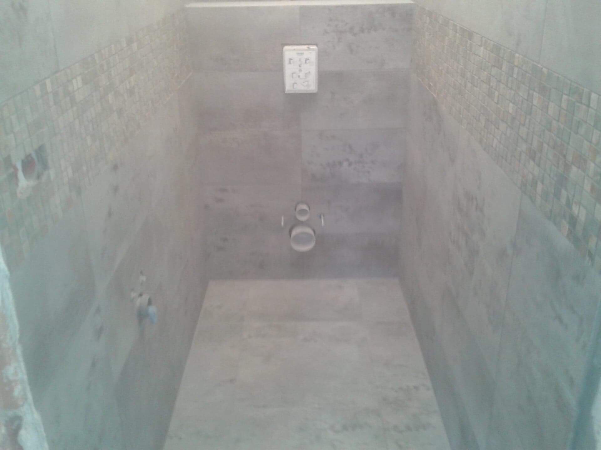 Wnętrze kabiny prysznicowej w trakcie wykańczania, widoczne szare płytki ceramiczne na ścianach i podłodze, dekoracyjny pas mozaiki na górze ściany, instalacja wodna oraz gniazdko elektryczne.