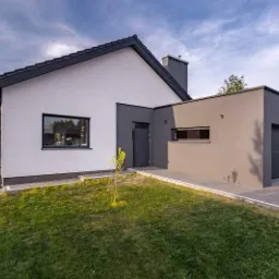 Nowoczesny dom jednorodzinny z białą elewacją, szarą dobudówką garażową i ciemnoszarą stolarką okienną, otoczony trawnikiem.