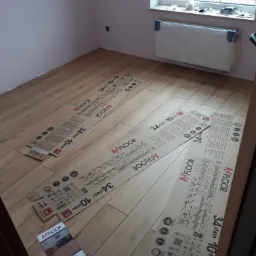 Przygotowanie do montażu paneli podłogowych Art Floor 34 class 10mm, rozłożone kartony z panelami na podłodze, widoczny grzejnik pod oknem.