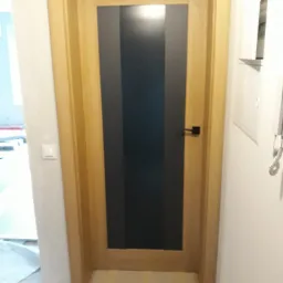 Zamknięte drzwi wewnętrzne z drewnianą ościeżnicą i czarnym, pionowym panelem pośrodku, widoczne zabezpieczenia podłogi taśmą malarską.