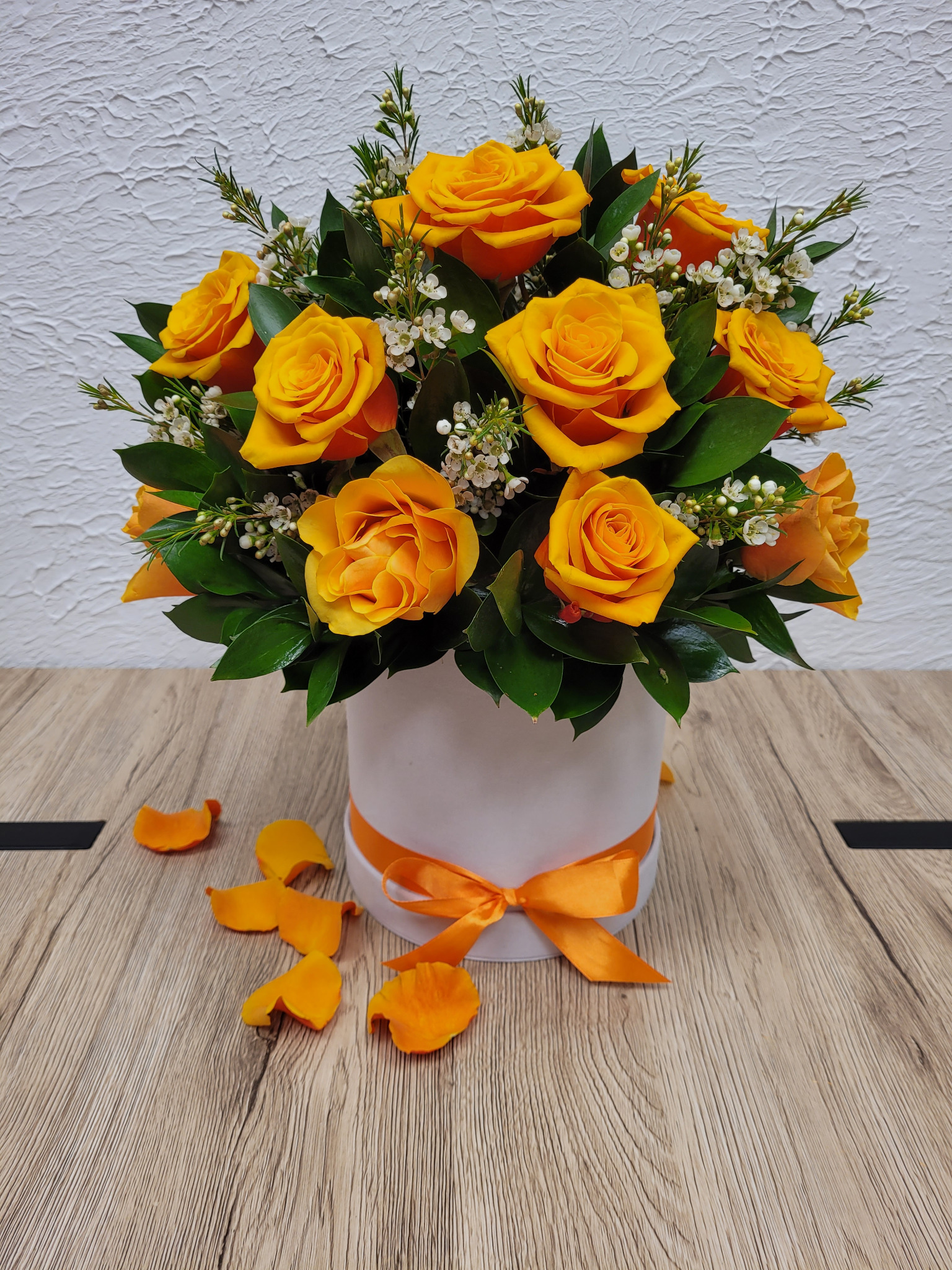 Kompozycja z herbacianych róż w białym, okrągłym pudełku, przewiązanym pomarańczową wstążką, na drewnianym blacie z kilkoma opadłymi płatkami róż.