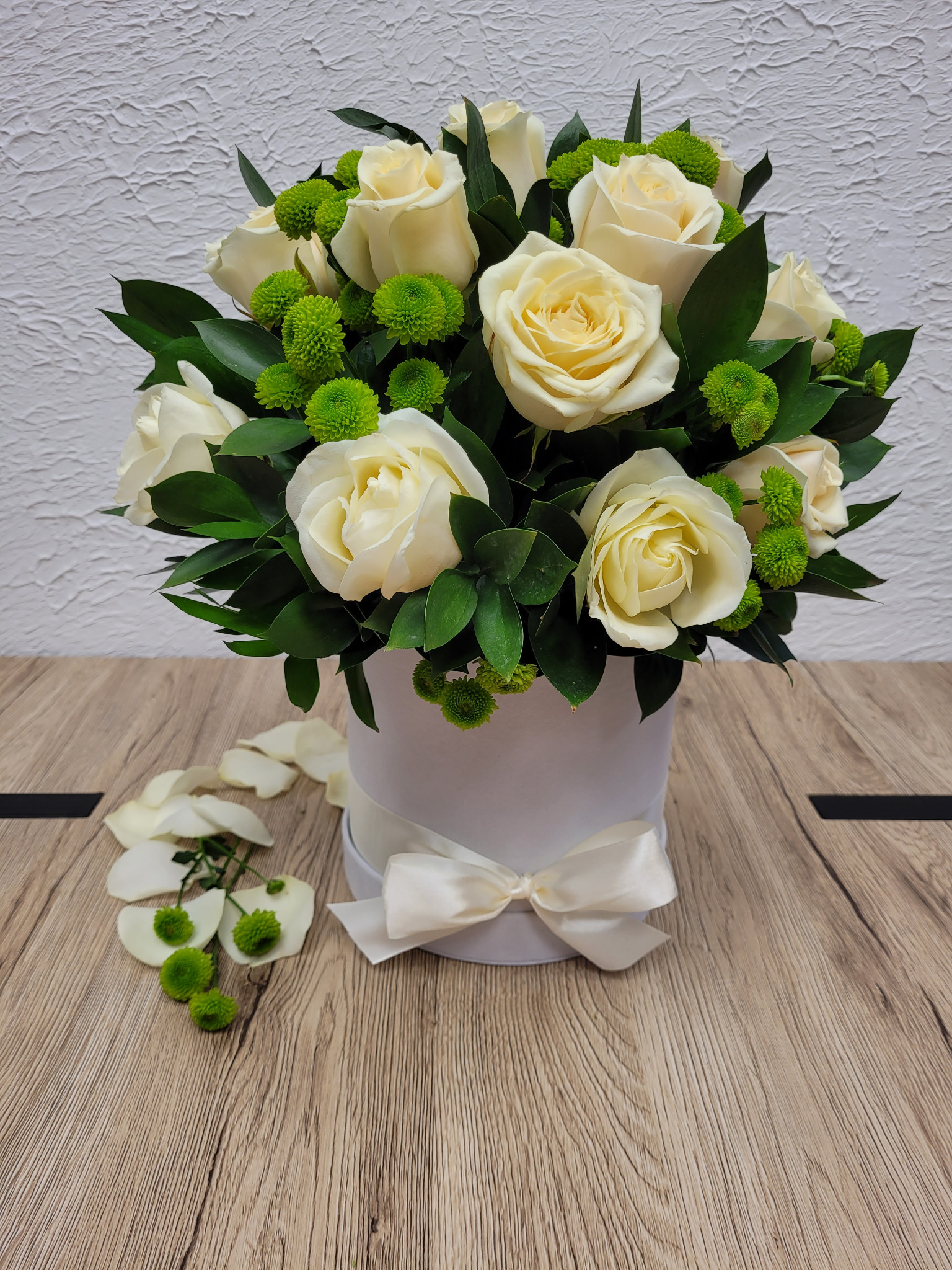 Kompozycja z białych róż i zielonych santini w białym pudełku z kokardą, leżąca na drewnianym blacie, z rozsypanymi płatkami róż i kwiatami santini obok.