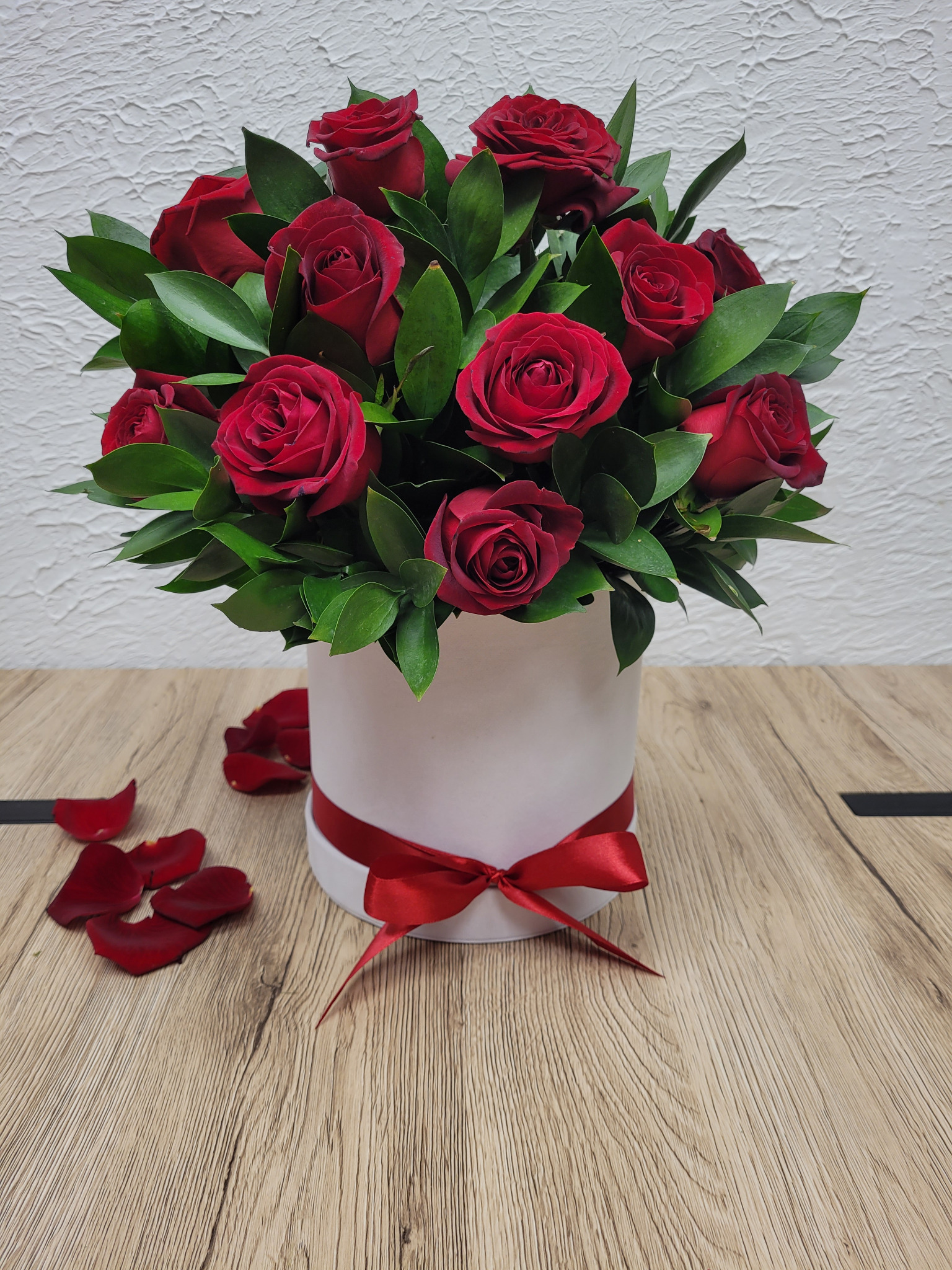 Bukiet czerwonych róż z zielonymi liśćmi w białym, okrągłym pudełku przewiązanym czerwoną wstążką, kilka płatków róż leży na drewnianym blacie.