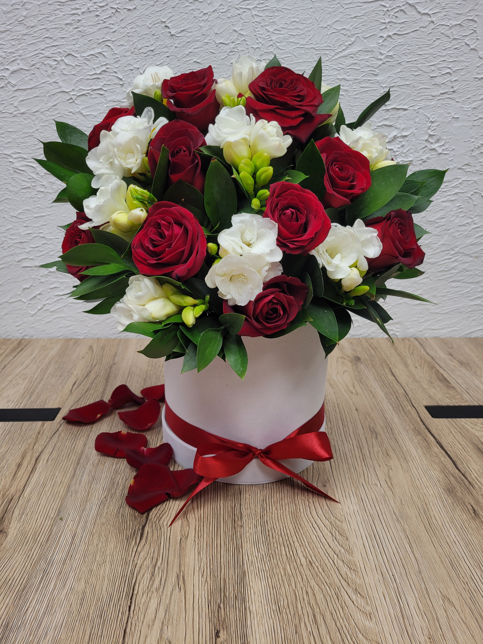 Elegancki bukiet z czerwonych róż i białych frezji w białym pudełku z czerwoną wstążką, leżący na drewnianym stole z rozrzuconymi płatkami róż.