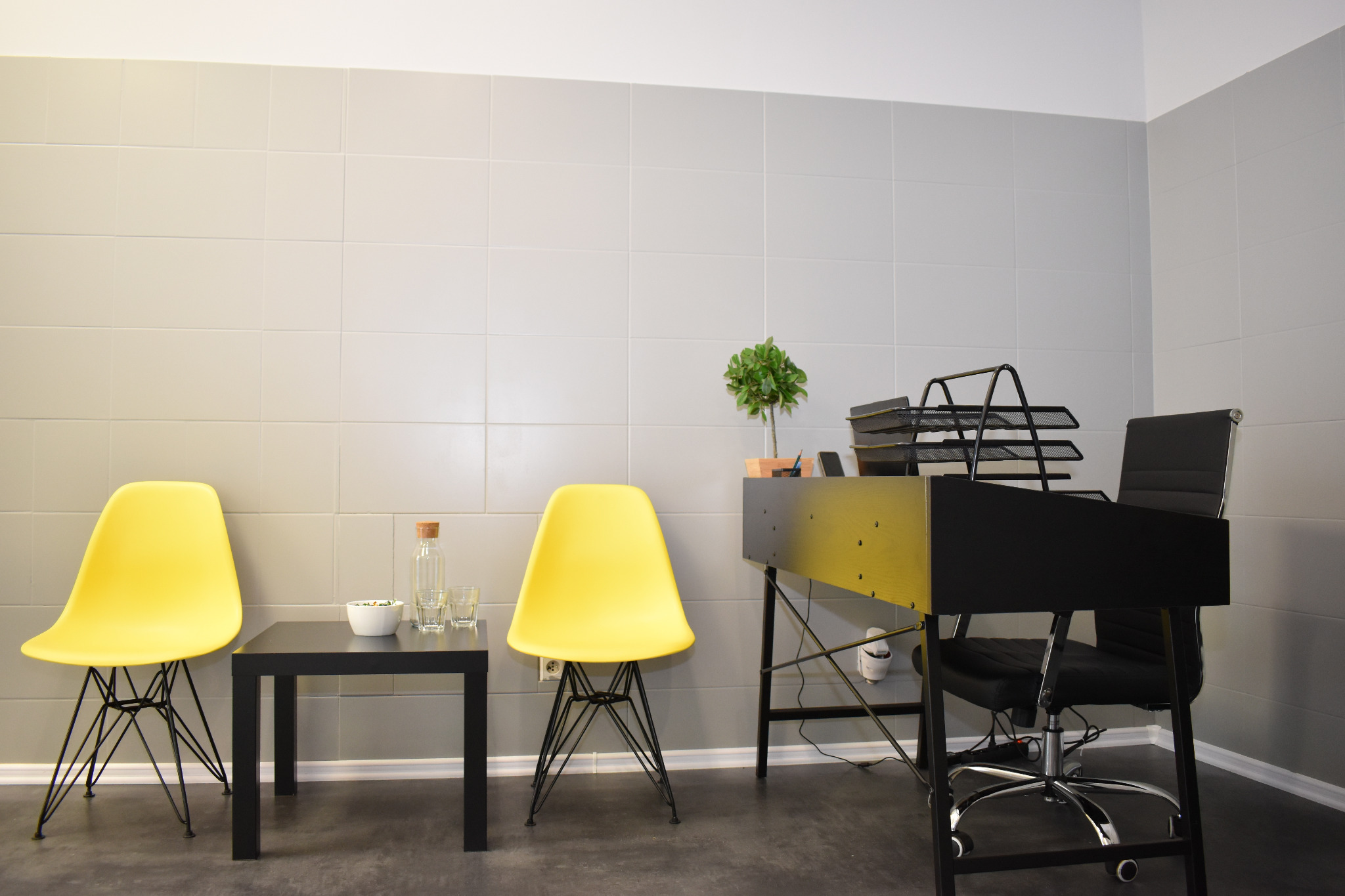 Nowoczesne biuro z dwoma żółtymi krzesłami, czarnym stolikiem z wodą i miską, oraz czarnym biurkiem z rośliną w doniczce i czarnym krzesłem biurowym na tle szarej ściany z płytek.