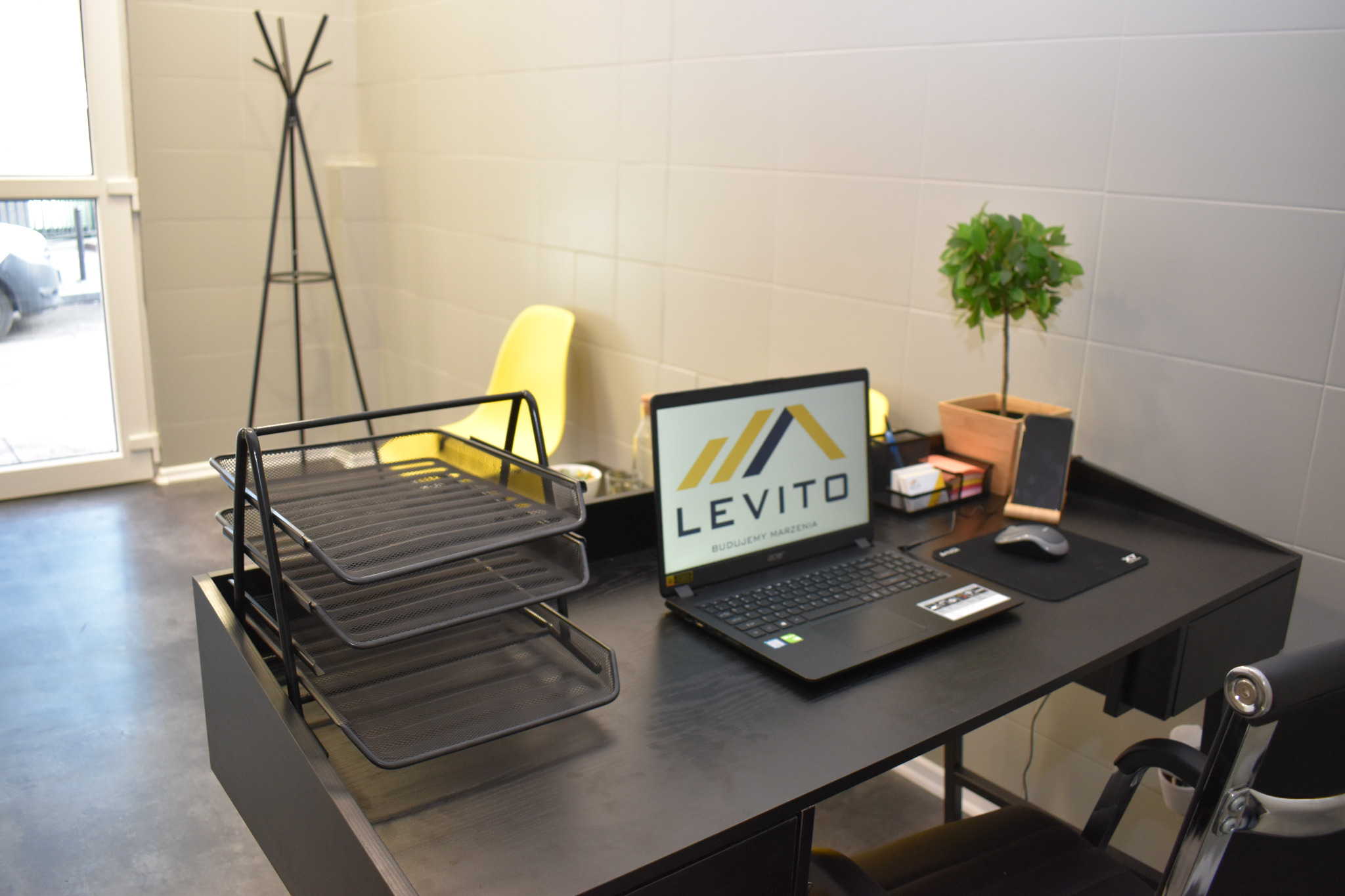 Biuro z czarnym biurkiem, laptopem z logo firmy budowlanej LEVITO, organizerem na dokumenty, małą rośliną doniczkową i żółtym krzesłem w tle.