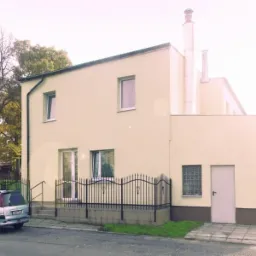 Dwukondygnacyjny budynek o jasnej elewacji z dwoma kominami, widoczny z ulicy, z zaparkowanym srebrnym kombi i ozdobnym metalowym ogrodzeniem.