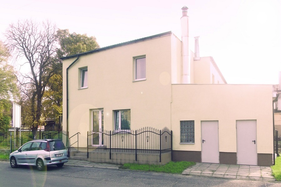 Dwukondygnacyjny budynek o jasnej elewacji z dwoma kominami, widoczny z ulicy, z zaparkowanym srebrnym kombi i ozdobnym metalowym ogrodzeniem.
