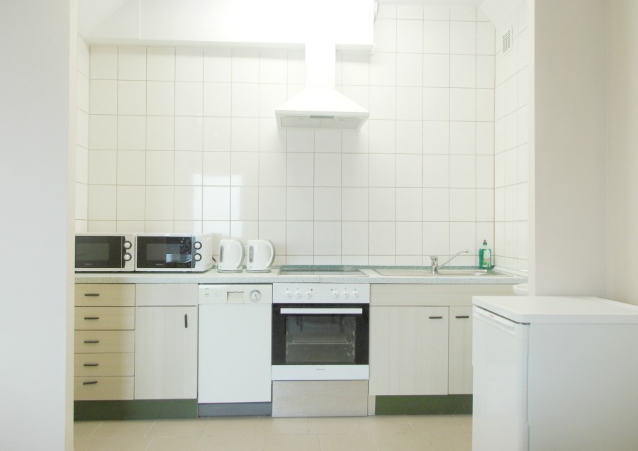 Jasna, minimalistyczna kuchnia z białymi płytkami, dwoma mikrofalówkami, czajnikami elektrycznymi, zlewem, okapem i lodówką, sprawiająca wrażenie czystej i funkcjonalnej.