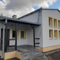 Elewacja budynku z białymi ścianami i rzędami okien z żółtymi obramowaniami, ciemne drzwi wejściowe pod zadaszeniem wspartym na czarnej drewnianej konstrukcji, szare schody wejściowe.