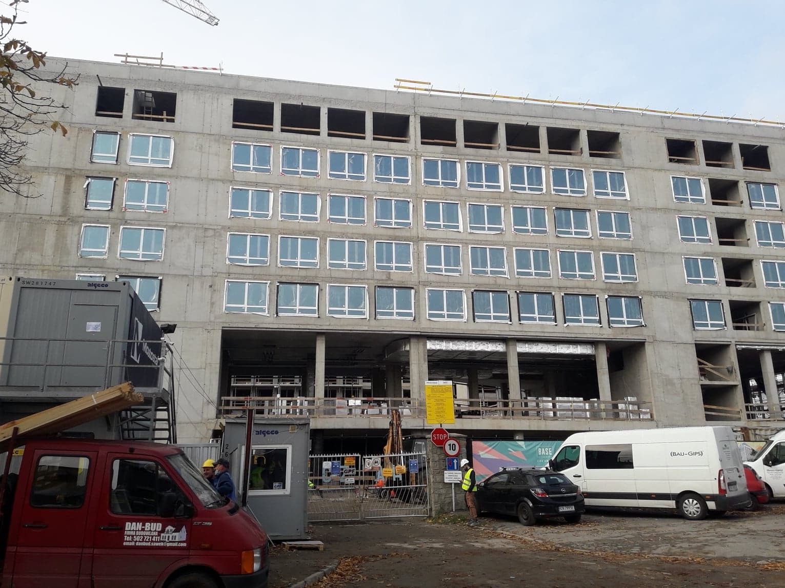 Wielopiętrowy budynek w trakcie montażu okien PCV, widoczne zabezpieczenia na oknach, plac budowy z kontenerami, samochodami dostawczymi i pracownikami w żółtych kaskach.