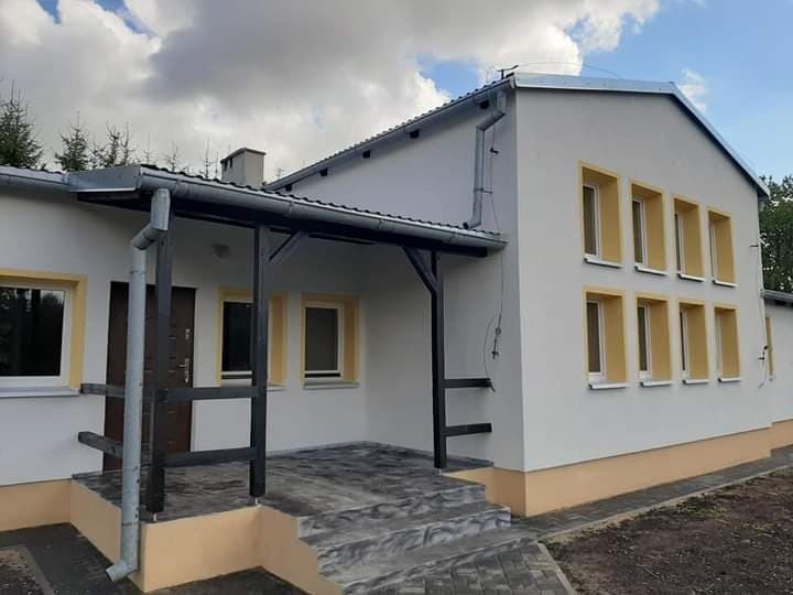 Elewacja budynku z białymi ścianami i rzędami okien z żółtymi obramowaniami, ciemne drzwi wejściowe pod zadaszeniem wspartym na czarnej drewnianej konstrukcji, szare schody wejściowe.