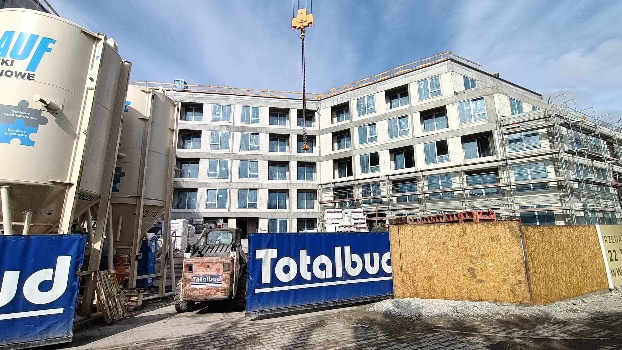 Budynek wielorodzinny w stanie surowym otwartym z zamontowanymi oknami, rusztowaniami i dźwigiem budowlanym, na pierwszym planie zbiorniki na materiały sypkie, miniładowarka i kontener z logo firmy...