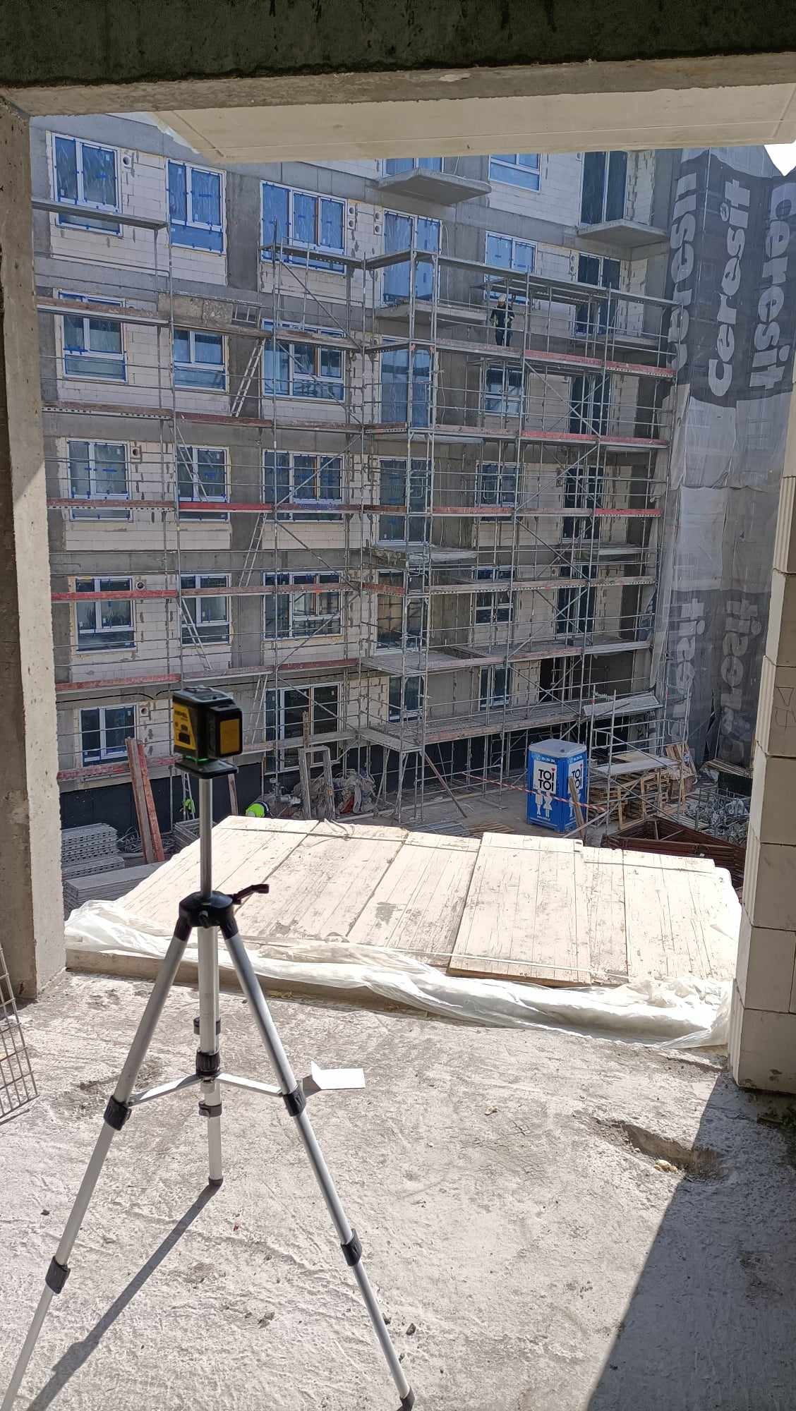 Widok z wnętrza budynku w trakcie budowy na rusztowania i wstawione okna w nowym budynku, z laserowym niwelatorem na statywie na pierwszym planie.
