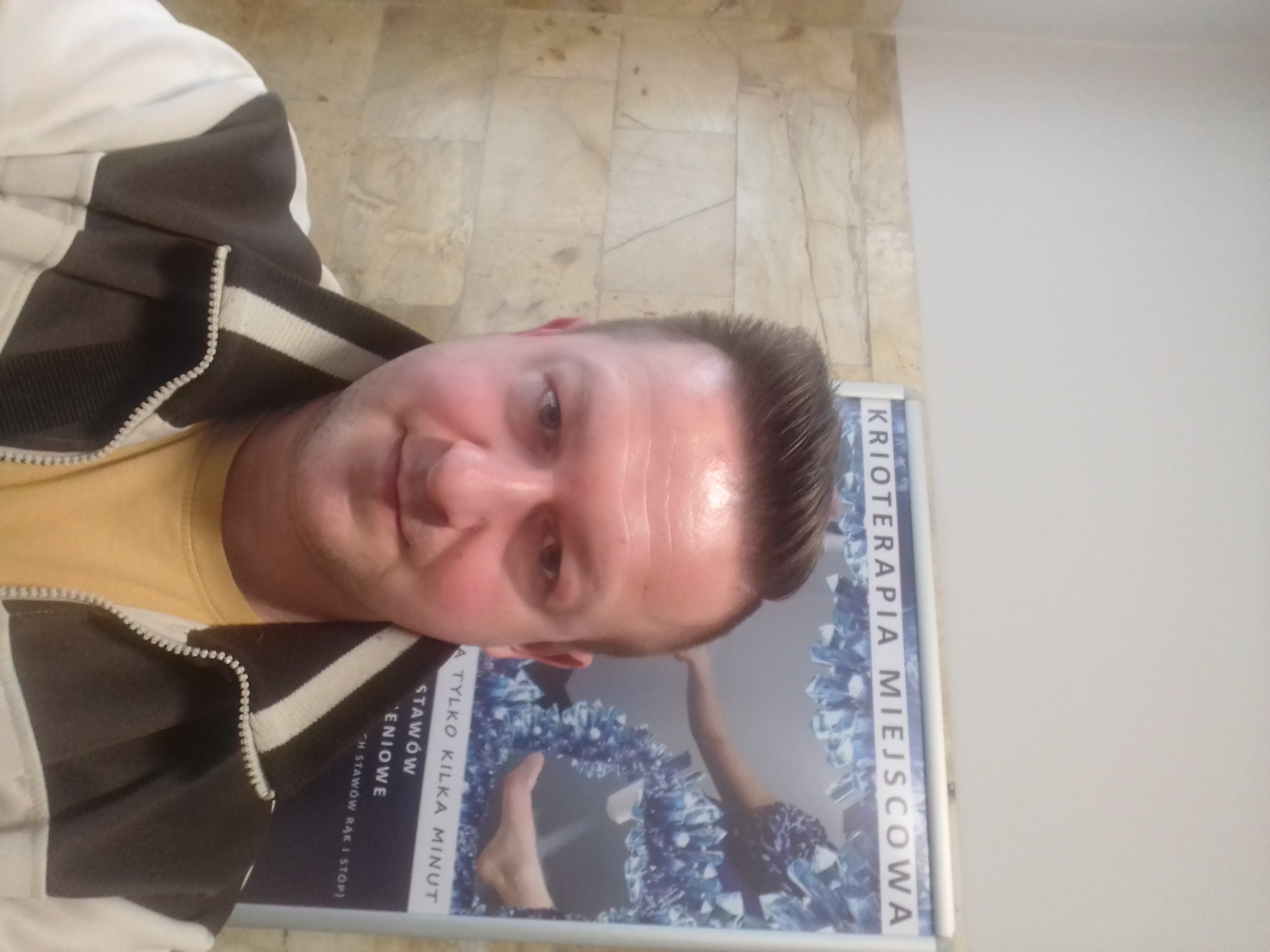 Mężczyzna robi selfie na tle plakatu reklamującego krioterapię miejscową, na plakacie widoczne są kryształki lodu i fragment ciała osoby poddawanej zabiegowi.