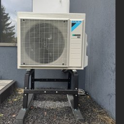 SUNLIT POMPY CIEPŁA DAIKIN, LG, HI-SENS, PANASONIC, PODŁOGÓWKA,REKUPERACJA, INSTALACJE GRZEWCZE - Zewnętrzna jednostka Daikin z widocznymi kroplami wody, zamontowana na czarnym metalowym stojaku, na tle szarej ściany budynku i okna odbijającego drzewo.