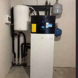 SUNLIT POMPY CIEPŁA DAIKIN, LG, HI-SENS, PANASONIC, PODŁOGÓWKA,REKUPERACJA, INSTALACJE GRZEWCZE - Kompletna instalacja grzewcza z białą pompą ciepła Daikin i zasobnikiem Galmet, z czarnymi izolowanymi rurami i dwoma zbiornikami wyrównawczymi, w narożniku pomieszczenia technicznego.
