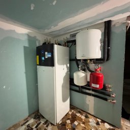 SUNLIT POMPY CIEPŁA DAIKIN, LG, HI-SENS, PANASONIC, PODŁOGÓWKA,REKUPERACJA, INSTALACJE GRZEWCZE - Zainstalowane białe urządzenie grzewcze z czarnym panelem i zasobnik c.w.u. Galmet, z rurami i zaworami, w pomieszczeniu z niedokończonymi ścianami i mozaikową podłogą.