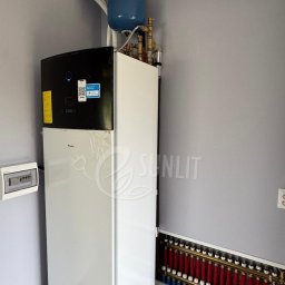 SUNLIT POMPY CIEPŁA DAIKIN, LG, HI-SENS, PANASONIC, PODŁOGÓWKA,REKUPERACJA, INSTALACJE GRZEWCZE - Nowoczesna, biała jednostka Daikin z czarnym panelem i niebieskim zbiornikiem, połączona z mosiężnymi rurami. Obok widoczny rozdzielacz do ogrzewania podłogowego w szarym pomieszczeniu technicznym.
