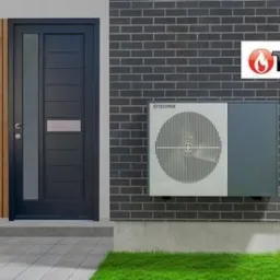 Zewnętrzna jednostka grzewcza zamontowana na ścianie budynku z ciemnej cegły, obok nowoczesnych drzwi wejściowych i drewnianej okładziny, widoczne logo producenta Technix.