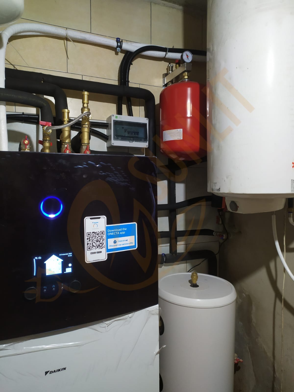Zainstalowana pompa ciepła Daikin z widocznym panelem sterowania, zbiornikiem buforowym i instalacją hydrauliczną w pomieszczeniu technicznym. Widoczny QR kod do aplikacji.