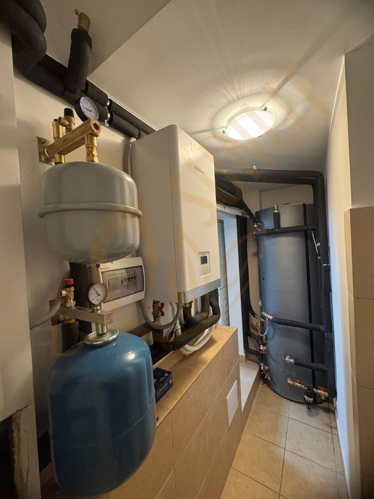 Wnętrze kotłowni z zainstalowanym kotłem grzewczym marki Kaisai, zbiornikiem akumulacyjnym i orurowaniem. Widoczne elementy instalacji i manometry.