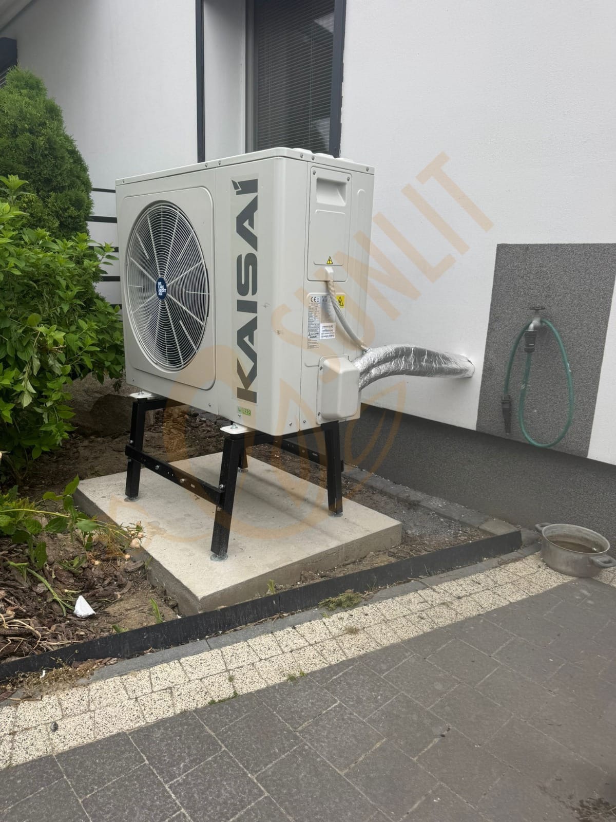 Biała pompa ciepła Kaisai zamontowana na czarnym stelażu, z podłączonymi przewodami, na tle białej ściany budynku z zielenią i elementami infrastruktury.