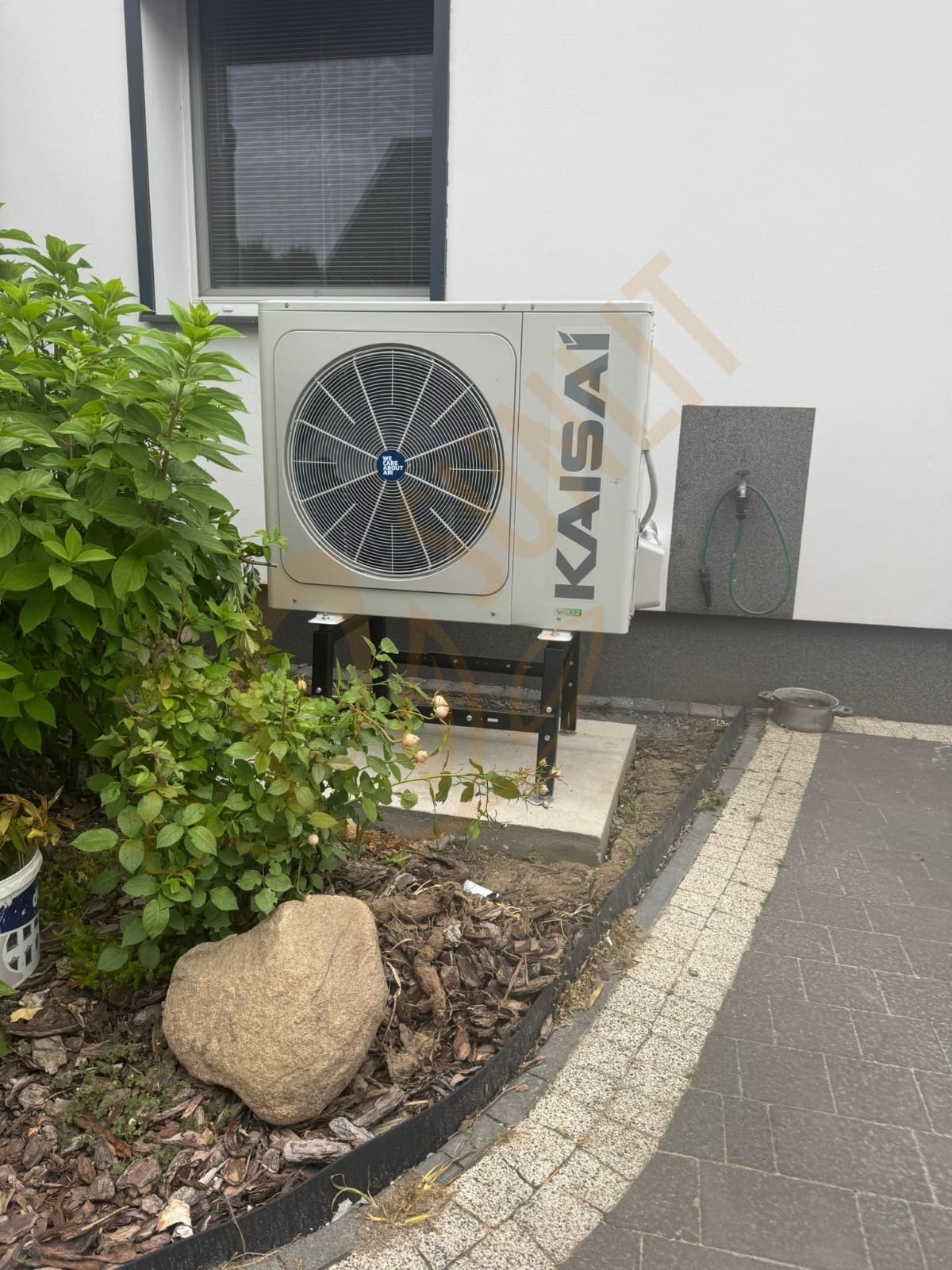 Szara pompa ciepła Kaisai zamontowana na zewnątrz budynku, widoczna z boku, z zielenią w tle i fragmentem chodnika z kostki brukowej.