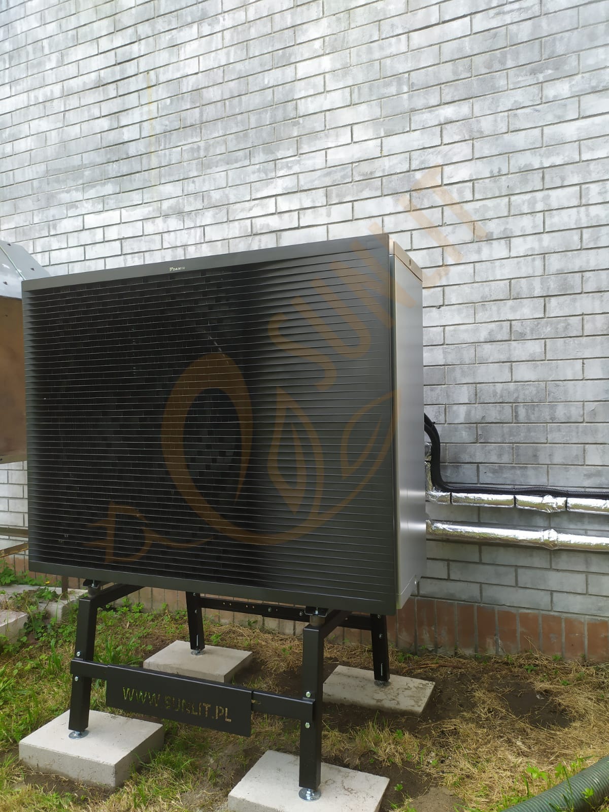 Czarna pompa ciepła Daikin na metalowym stelażu, ustawiona na betonowych płytach przed ceglaną ścianą. Widoczne rury w izolacji. Na pompie logo firmy w formie grafiki.