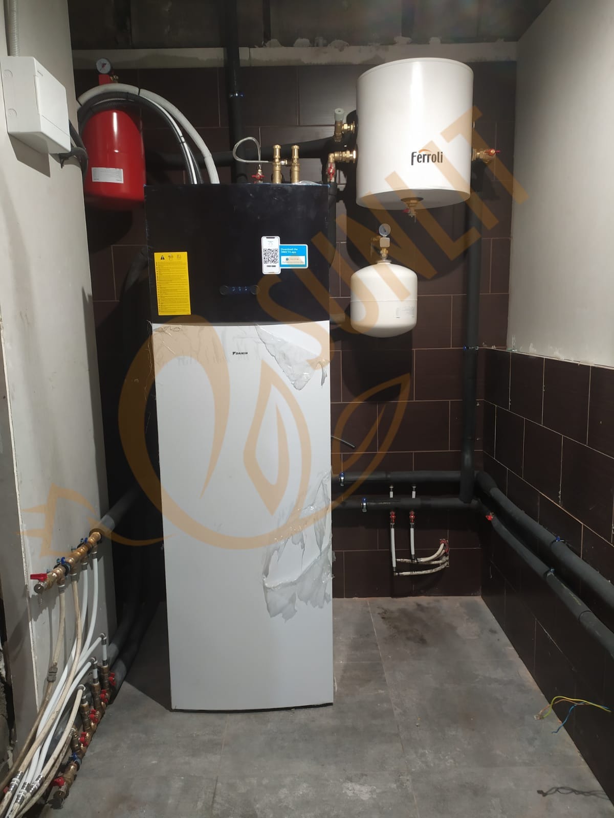 Instalacja pompy ciepła Daikin z osprzętem Ferroli w kotłowni z brązowymi płytkami. Widoczne rury i zbiorniki. Część pompy zabezpieczona folią.
