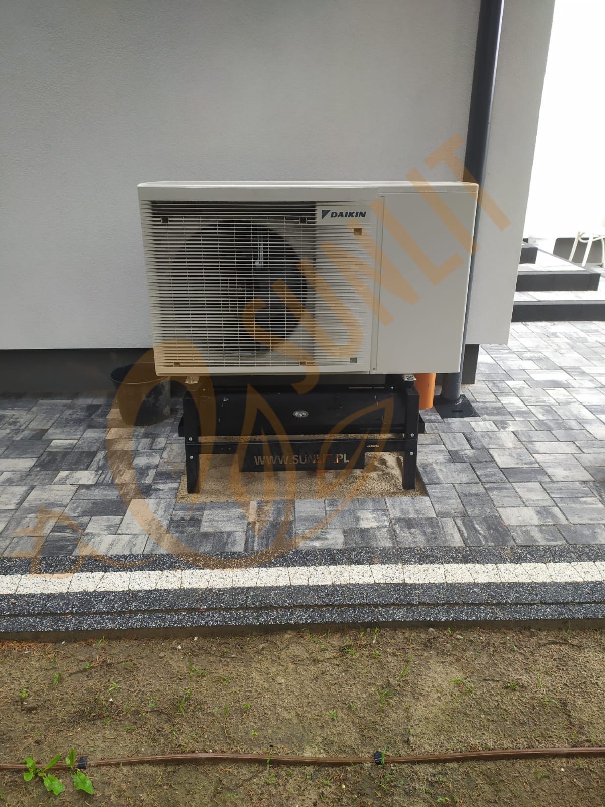 Biała pompa ciepła Daikin zamontowana na czarnym stelażu na kostce brukowej. Widoczny fragment elewacji budynku i trawnika z systemem nawadniania.