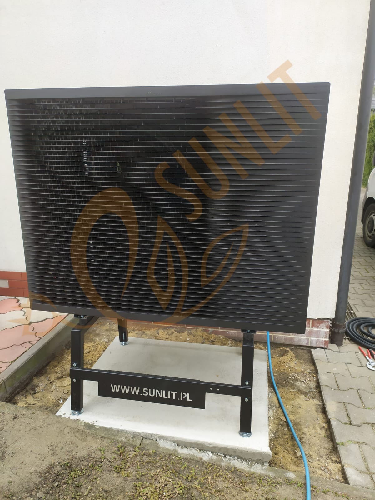 Czarna pompa ciepła marki Sunlit zamontowana na betonowej podstawie przy ścianie budynku. Widoczne przewody i narzędzia.