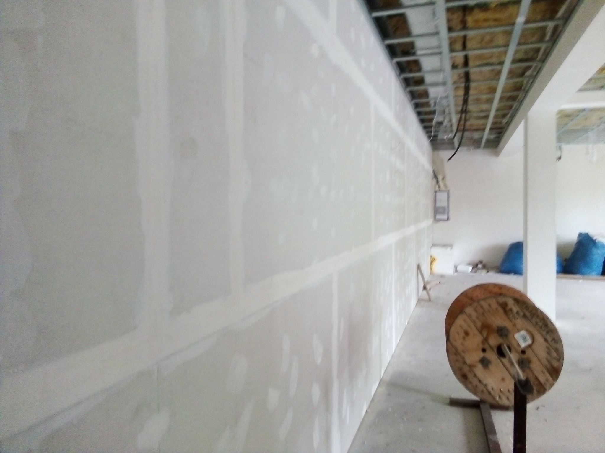 Ściana z płyt gipsowo-kartonowych po nałożeniu pierwszej warstwy masy szpachlowej, widoczna konstrukcja sufitu podwieszanego i pusta szpula po kablu w pomieszczeniu w trakcie remontu.