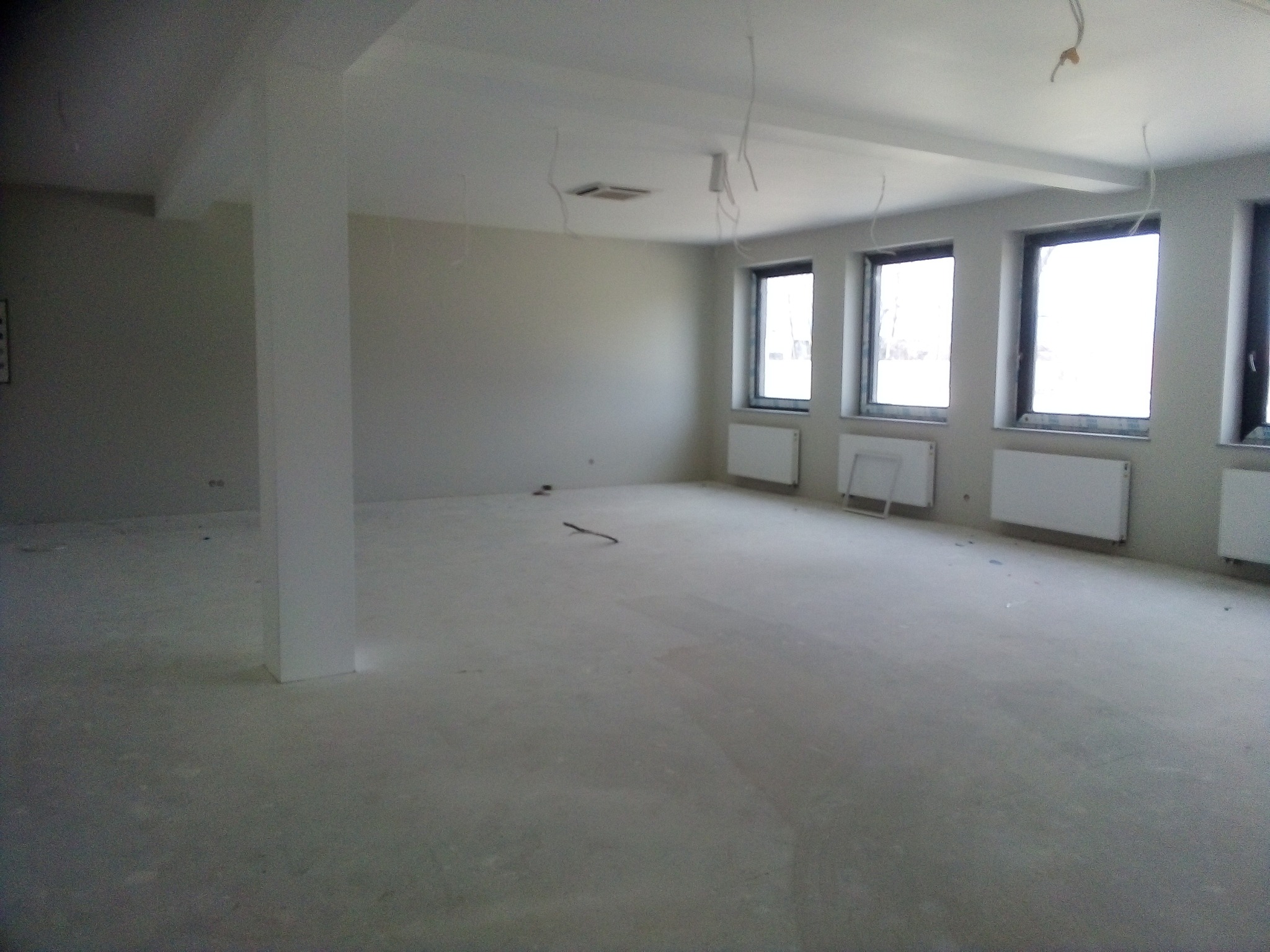 Duże, puste pomieszczenie w stanie surowym z widocznymi grzejnikami pod oknami i wystającymi przewodami elektrycznymi z sufitu, filarem konstrukcyjnym po lewej stronie.