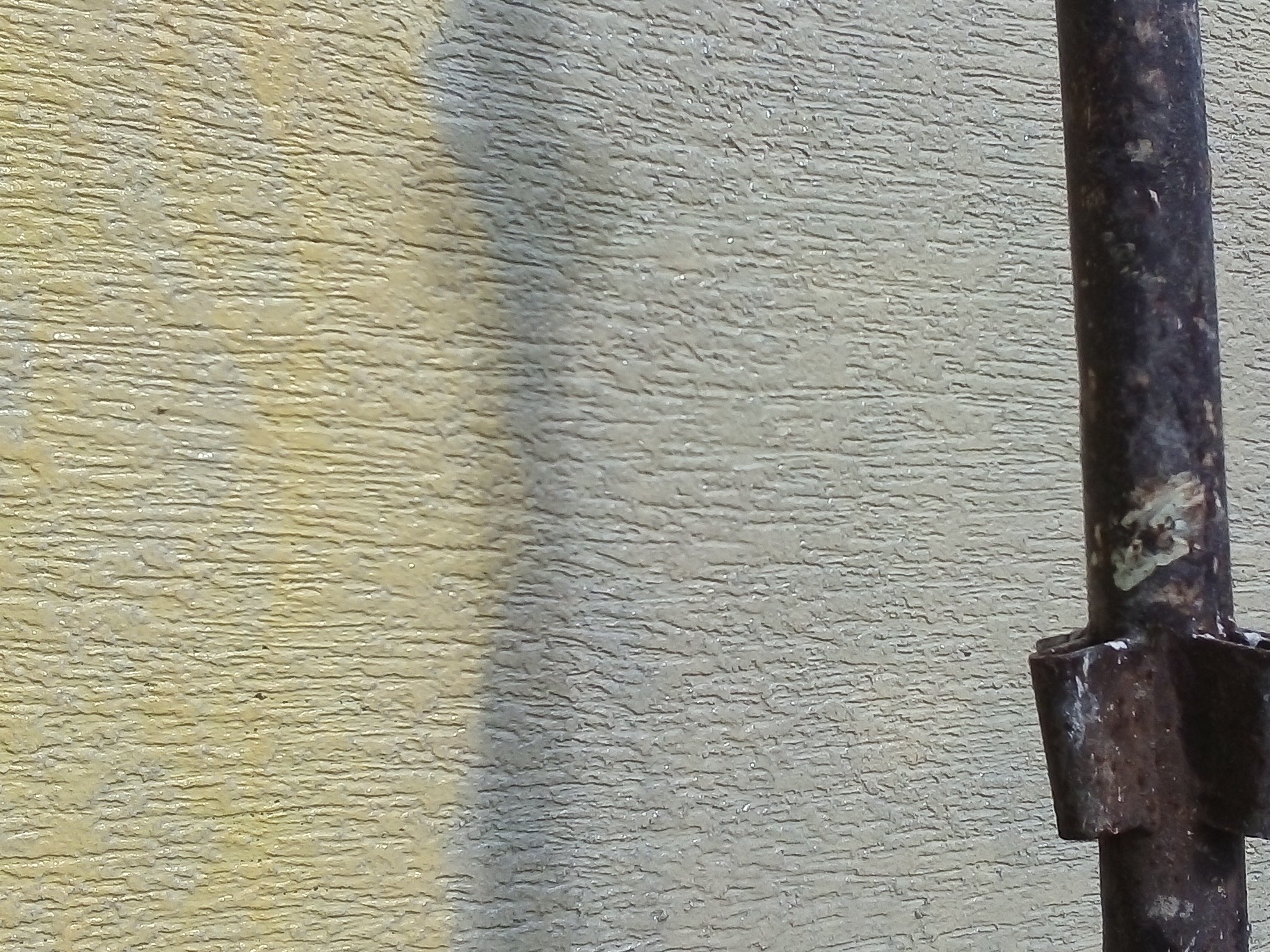 Fragment teksturowanej ściany w odcieniach beżu i szarości, z fragmentem zardzewiałej metalowej rury po prawej stronie kadru.