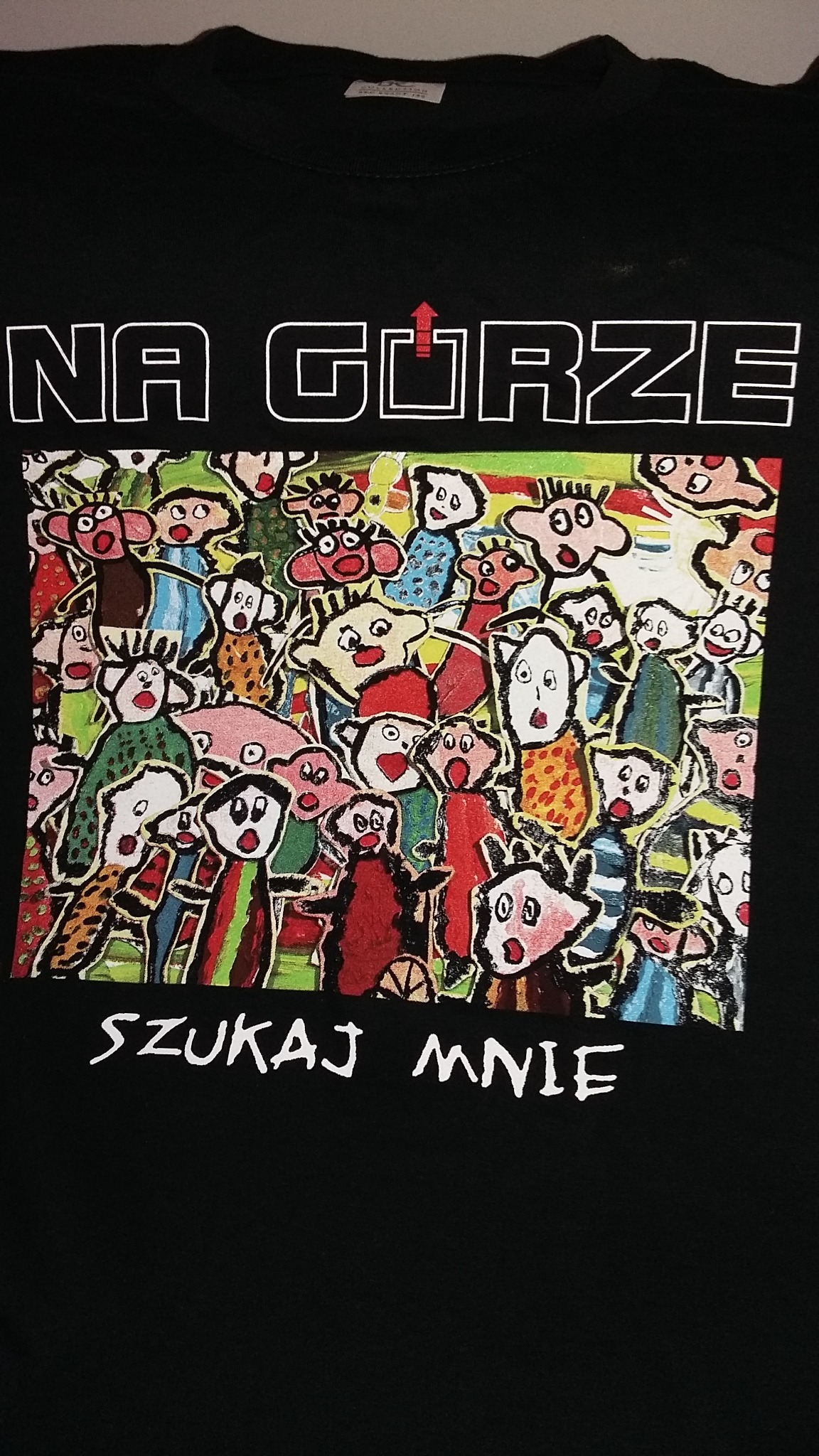 Czarna koszulka z kolorowym, ręcznie rysowanym nadrukiem przedstawiającym tłum postaci i napisy 'NA GÓRZE' oraz 'SZUKAJ MNIE'.