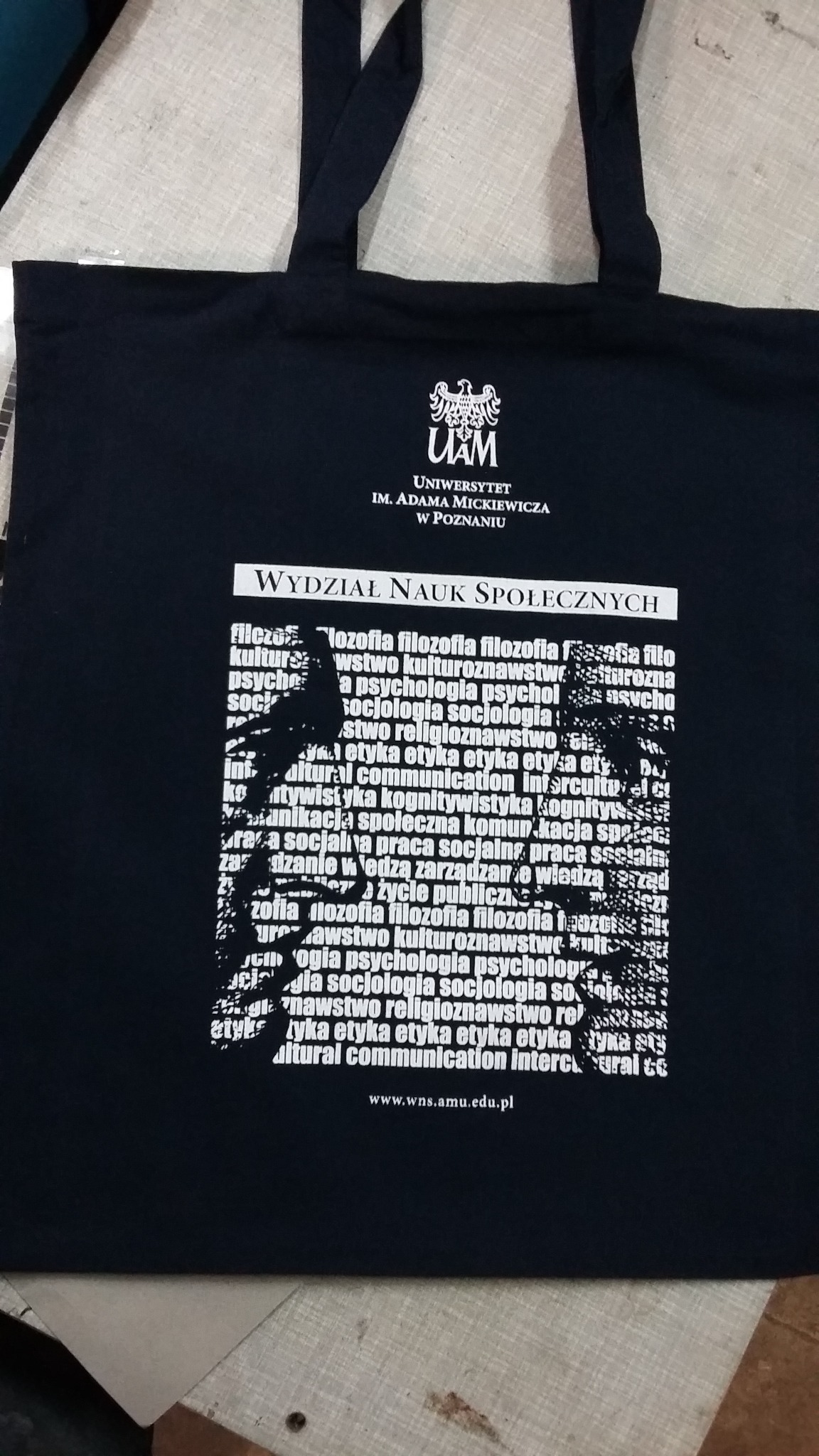 Czarna torba bawełniana z nadrukiem logo Uniwersytetu im. Adama Mickiewicza w Poznaniu i nazw Wydziału Nauk Społecznych wraz z listą kierunków studiów.