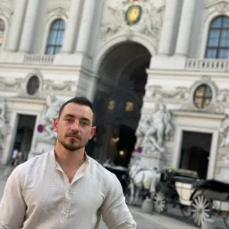 Mężczyzna w lnianej koszuli na tle Hofburga w Wiedniu, z dorożkami konnymi w tle.