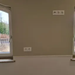 Ściana w odcieniach beżu z dwoma gniazdkami elektrycznymi i jednym potrójnym gniazdem antenowym, widok z okna na zewnątrz.