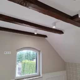 Poddasze z drewnianymi belkami stropowymi, trzema okrągłymi lampami punktowymi, oknem w kształcie łuku, gniazdkami elektrycznymi i tapetą w kratę.