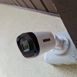 Kamera monitoringu marki KONIK zamontowana na jasnej elewacji pod drewnianym okapem, widok z dołu.