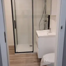 Nowoczesna łazienka z kabiną prysznicową z czarnymi ramami, białą umywalką z szafką i toaletą jeszcze w folii ochronnej, wykończona panelami podłogowymi imitującymi drewno.