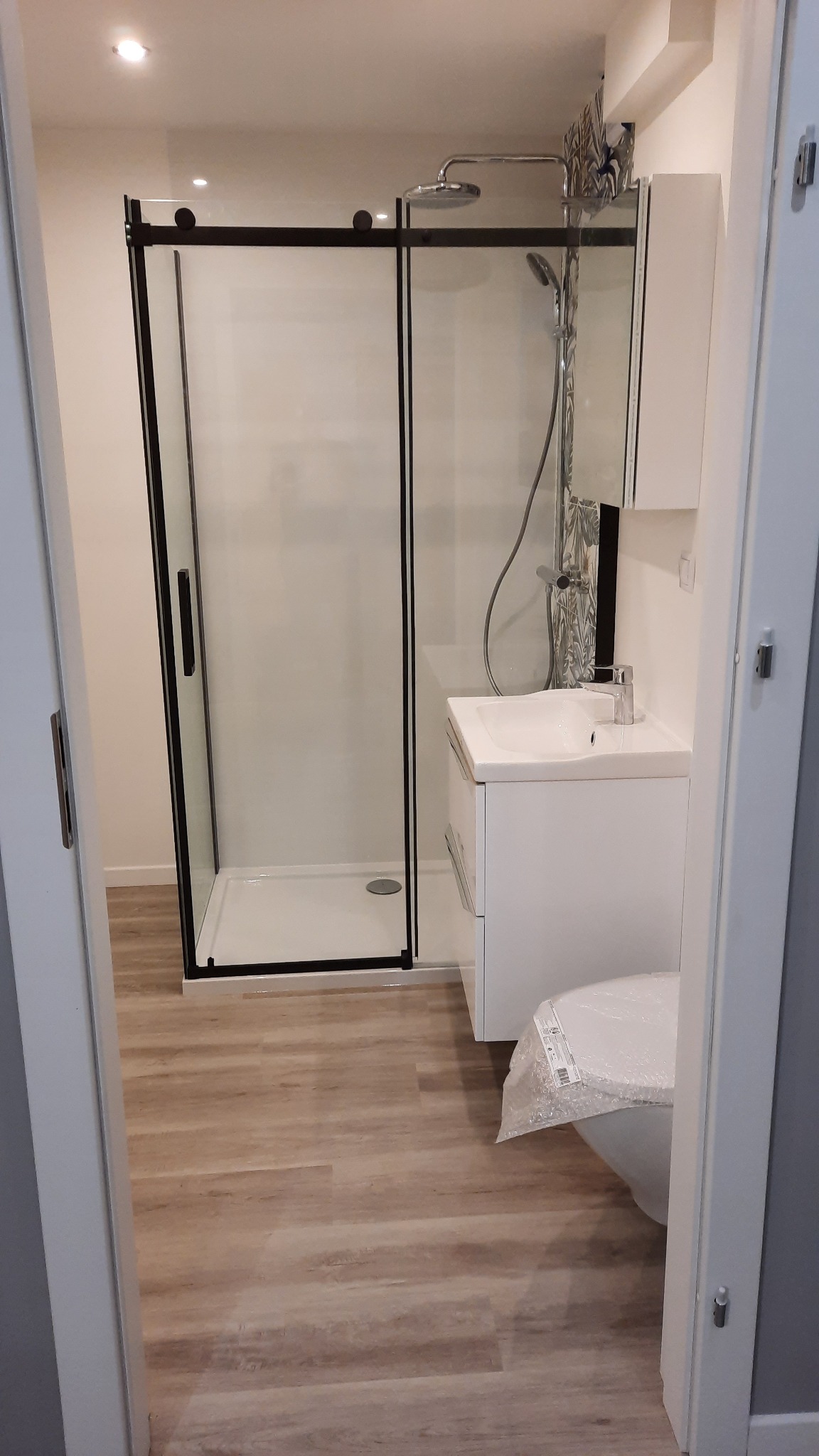 Nowoczesna łazienka z kabiną prysznicową z czarnymi ramami, białą umywalką z szafką i toaletą jeszcze w folii ochronnej, wykończona panelami podłogowymi imitującymi drewno.