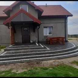 Szary dom z czerwonym dachem i nowymi schodami wejściowymi z kostki brukowej w odcieniach szarości, z drewnianą ławką po prawej stronie.