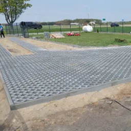 Układanie szarej ażurowej kostki brukowej na piaskowym podłożu, widoczny fragment ułożonej powierzchni, w tle trawnik, drzewa, ogrodzenie i zabudowania.