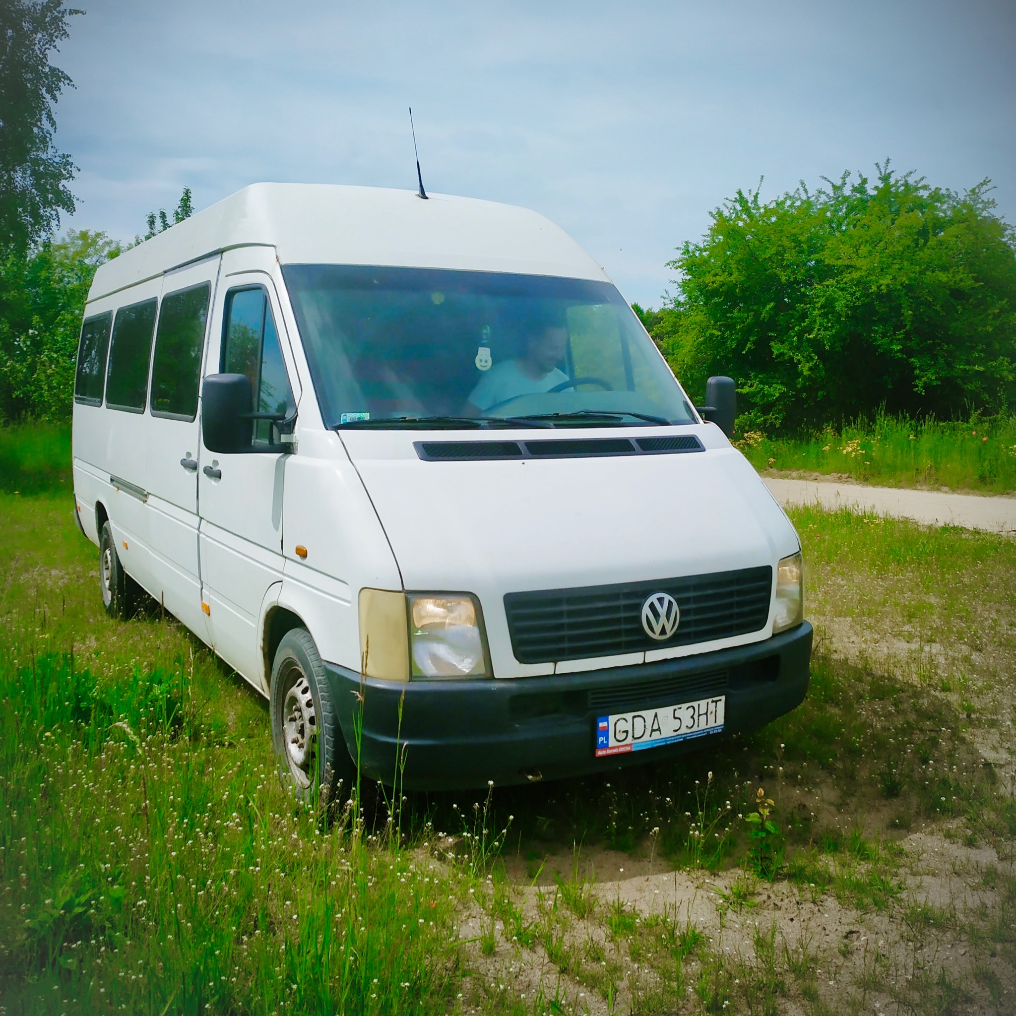 Biały bus Volkswagen LT z polską tablicą rejestracyjną GDA 53HT, zaparkowany na piaszczystej drodze zarośniętej trawą, z widocznym kierowcą w środku.
