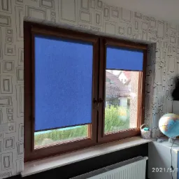 Okno z brązowymi ramami, na którym zamontowano niebieskie rolety w kasecie, widoczne z zewnątrz zielone krzewy i domy, w tle ściana z tapetą w geometryczne wzory.