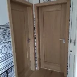 Dwa drewniane drzwi wewnętrzne z prostokątnymi panelami, uchwyty w kolorze srebrnym, widoczne na tle ściany z czarno-białą tapetą przedstawiającą architekturę miejską i drewnianą podłogą.